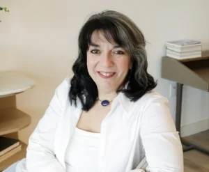margaret warner wearing a white camisole and white button down sitting on a chair